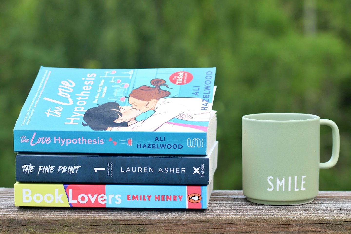 Three romance novels and a mug on a wooden railing.